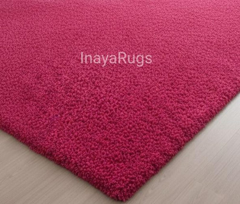 May include: A close-up of a textured, magenta-colored rug. The rug has a dense, plush pile and the text "Inaya Rugs" is visible. The rug's corner is visible, showing its thick construction.