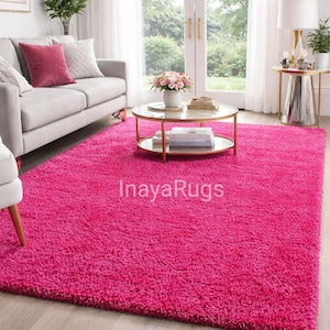 May include: A vibrant pink shag rug dominates the room, covering the floor. The rug is rectangular and plush, with a deep pile. The room also features a gray sofa with decorative pillows, a round coffee table, and a side table with a lamp. The text "InayaRugs" is visible on the rug.