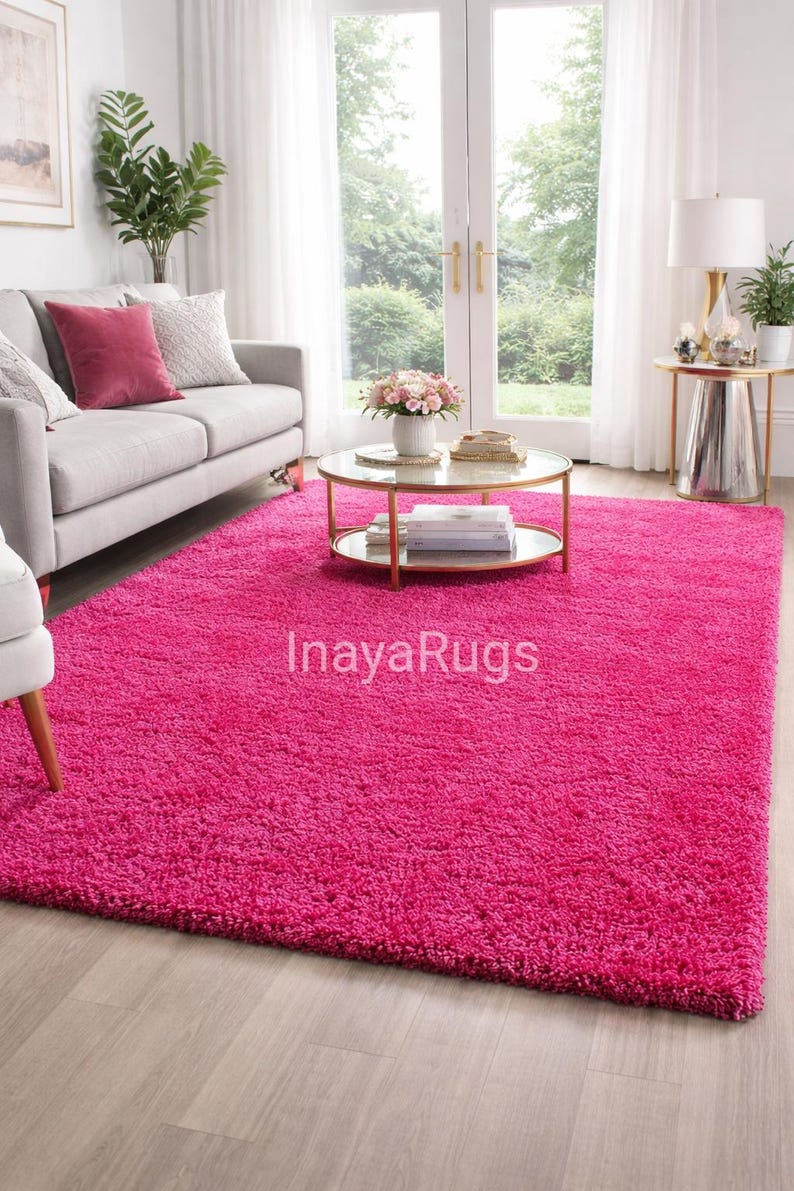 May include: A vibrant pink shag rug dominates the room, covering the floor. The rug is rectangular and plush, with a deep pile. The room also features a gray sofa with decorative pillows, a round coffee table, and a side table with a lamp. The text "InayaRugs" is visible on the rug.