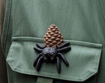 May include: A black spider and pinecone brooch pinned to a green vest pocket. The spider has eight legs and is positioned below the brown pinecone. The vest is a solid green color.