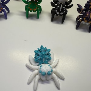Peut inclure: Collection de figurines d'araignées colorées imprimées en 3D. Les araignées sont de différentes couleurs, dont le blanc, le bleu, le vert, le violet, le noir et le jaune. Chaque araignée a un design unique, avec des détails et des textures. Les figurines sont disposées sur une surface blanche.