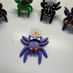 Peut inclure: Plusieurs figurines d'araignées imprimées en 3D aux couleurs vives. Les araignées présentent différents motifs, dont une citrouille, un crâne et une fleur de lotus. Les figurines sont dans des tons d'orange, de vert, de noir, de blanc et de violet. Les araignées ont plusieurs pattes.