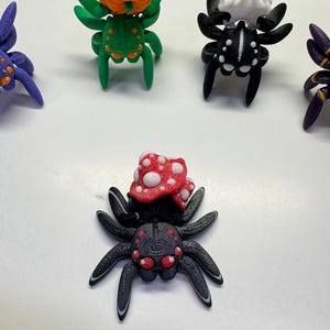 Peut inclure: Plusieurs figurines d'araignées imprimées en 3D. Une araignée verte porte une citrouille orange sur son dos. Une autre araignée noire a un crâne blanc. Une troisième araignée noire a un champignon rouge et blanc.