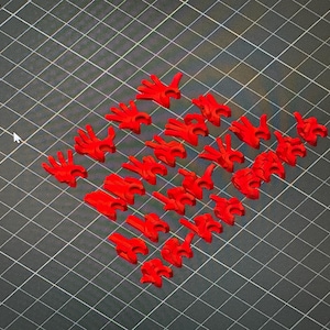 Może przedstawiać: Zbiór małych, jaskrawoczerwonych, drukowanych w 3D, przegubowych dłoni i elementów ramion, ułożonych na czarnej powierzchni kratki. Części różnią się rozmiarem i pozą, prawdopodobnie do projektu montażu modelu.