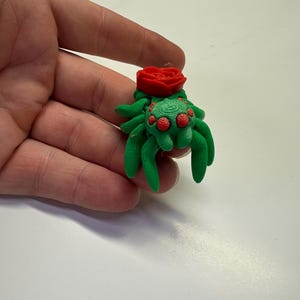 Peut inclure: Figurine décorative d'araignée verte et rouge avec une rose rouge sur le dos. L'araignée a plusieurs yeux rouges et est tenue dans une main. La figurine est probablement en plastique ou en argile.