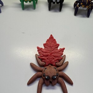 Peut inclure: Figurine d'araignée marron et rouge avec un motif en forme de feuille sur le dos. L'araignée a plusieurs yeux et des pattes détaillées. D'autres figurines d'araignées colorées sont visibles en arrière-plan.