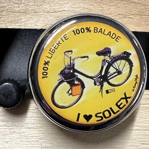 May include: A circular badge with a silver edge and a yellow centre, depicting a black bicycle. The text reads "100% LIBERTE" and "100% BALADE", with "I ❤️ SOLEX" at the bottom. A black bottle cap is visible.