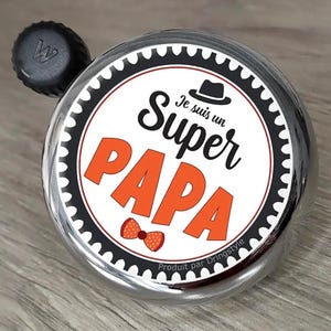 Peut inclure: Une sonnette de vélo ronde en argent avec un percuteur noir, dotée d'une étiquette circulaire blanche. L'étiquette affiche une icône de chapeau fedora noir, le texte 'Je suis un Super PAPA' en noir et orange, et un petit nœud papillon orange à pois.