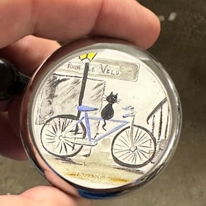 Op de afbeelding: Een zilveren fietsbel met een handgeschilderd ontwerp. De afbeelding toont een zwarte kat op een blauwe fiets, met een straatlantaarn en de tekst "Pour le Velo". De bel heeft een glanzend, reflecterend oppervlak.