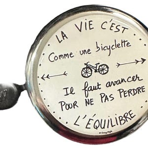Peut inclure: Une sonnette de vélo argentée avec un cadran blanc et des lettres noires. La sonnette porte l'inscription "LA VIE C'EST Comme une bicyclette Il faut avancer POUR NE PAS PERDRE L'EQUILIBRE".