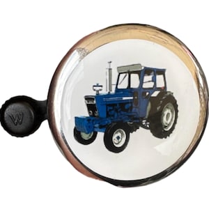 Op de afbeelding: Een ronde, zilverkleurige bel met een blauw tractormotief. De bel heeft een zwarte handgreep met de letter "W" in reliëf. De tractor is blauw met witte banden en een wit dak.