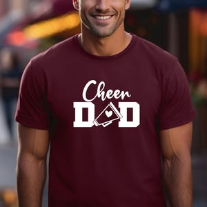 May include: A maroon t-shirt with white text that reads "Cheer Dad" with a white megaphone and heart graphic.