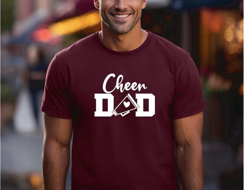 May include: A maroon t-shirt with white text that reads "Cheer Dad" with a white megaphone and heart graphic.