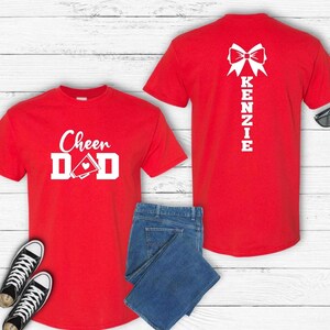 May include: Two red t-shirts with white text and graphics. The front shirt says "Cheer Dad" with a megaphone and heart. The back shirt has a bow and the name "Kenzie" written vertically.