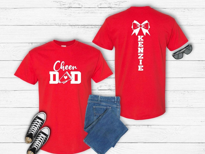 May include: Two red t-shirts with white text and graphics. The front shirt says "Cheer Dad" with a megaphone and heart. The back shirt has a bow and the name "Kenzie" written vertically.