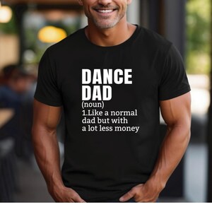 May include: Black t-shirt with the text "DANCE DAD" in white, followed by a definition. The definition reads: "1. Like a normal dad but with a lot less money."