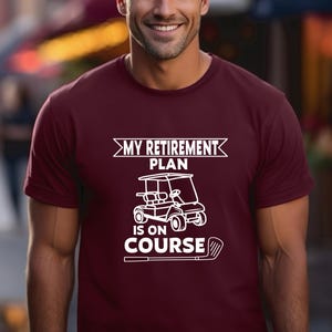 K&ouml;nnte beinhalten: Ein maroonfarbenes T-Shirt mit einer wei&szlig;en Grafik eines Golfwagens und dem Text "My Retirement Plan Is On Course".