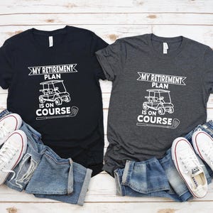K&ouml;nnte beinhalten: Zwei T-Shirts mit einem Golfwagen-Motiv und dem Text "My Retirement Plan Is On Course". Ein Shirt ist schwarz und das andere dunkelgrau.