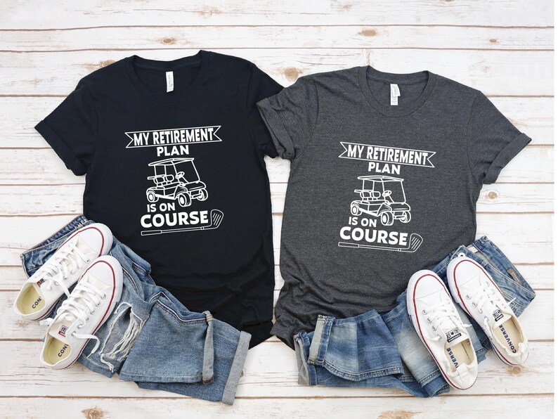 K&ouml;nnte beinhalten: Zwei T-Shirts mit einem Golfwagen-Motiv und dem Text "My Retirement Plan Is On Course". Ein Shirt ist schwarz und das andere dunkelgrau.