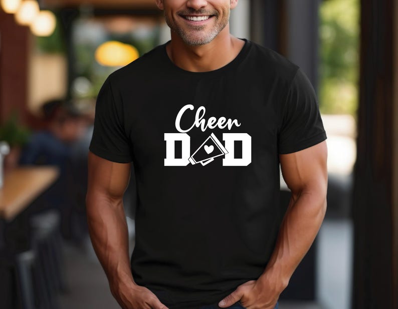 May include: Black t-shirt with white text that reads "Cheer Dad" and a white megaphone with a heart.