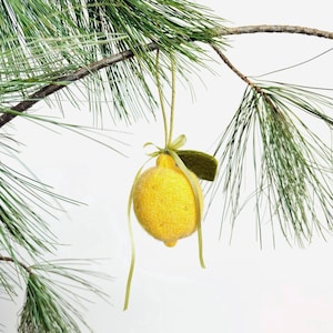 May include: A yellow knitted lemon ornament with a green leaf and ribbon, suspended from a pine branch. The lemon has a textured surface and is tied with a green ribbon. A festive, handcrafted decoration.