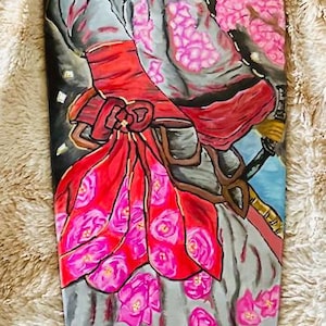 May include: A blue and white skateboard deck with a hand-painted design of a person in a red and pink kimono with a green headband. The person is holding a sword and is surrounded by pink flowers. The word "ENSO" is written in white on the bottom of the deck.
