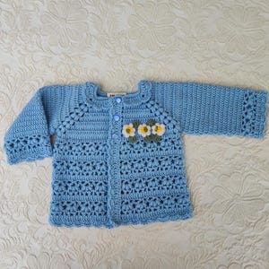 May include: A light blue crocheted baby cardigan with a white collar and button closure. The cardigan has a floral design on the front with white and yellow flowers.