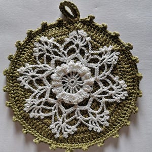 May include: A handmade, round crochet potholder in olive green with a white floral design in the centre. The potholder has a loop for hanging and a scalloped edge. The design features a large white flower in the centre.