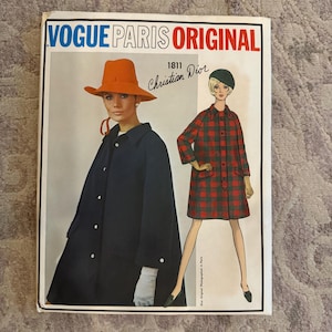May include: A vintage Vogue Paris Original sewing pattern featuring a Christian Dior design. The cover showcases a woman in a navy coat and orange hat, and another in a red and green plaid coat. The text reads "Vogue Paris Original 1811 Christian Dior."
