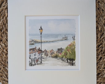May include: A watercolor painting in a white mat board featuring a coastal town scene with a steep street leading toward the sea. The artwork depicts white houses with red roofs, a tall black lamppost, and green foliage overlooking a harbor.
