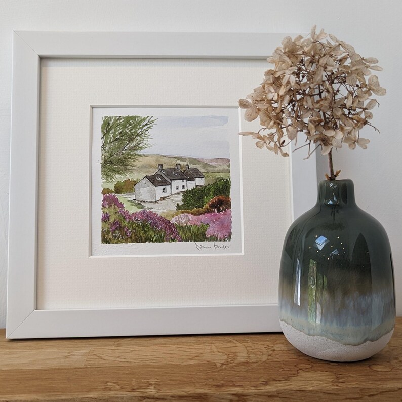 May include: A framed watercolor painting of a white cottage with a green roof, surrounded by purple flowers and green trees. The painting is signed "K.Anne Eyre" in the bottom right corner.