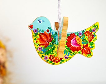 May include: A wooden bird ornament with a light blue head and yellow body, adorned with vibrant floral patterns in red, orange, and purple. The bird hangs from a silver chain, showcasing a folk art style.
