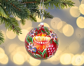 May include: A red Christmas bauble decorated with a floral pattern in white, blue, and orange. The bauble hangs from a silver string, set against a blurred background of lights and a green pine branch.