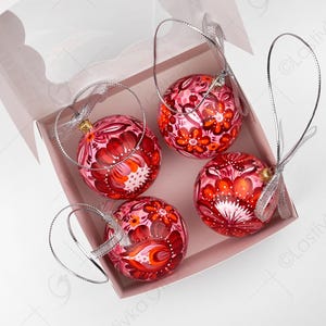 May include: Four hand-painted Christmas ornaments in a pink box. The ornaments are round, with a red and pink floral design. Each ornament has a silver ribbon for hanging. The box is light pink.