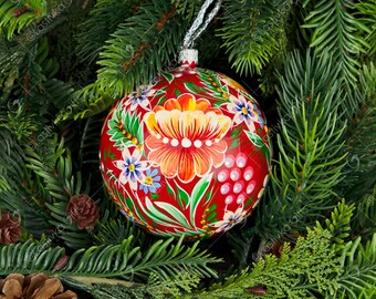May include: A red Christmas bauble with a floral design painted on it. The bauble is surrounded by green pine branches.