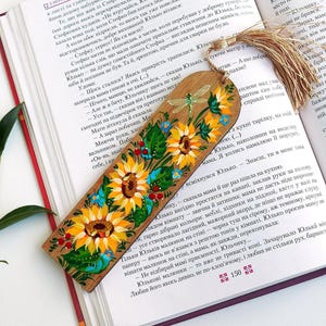 Op de afbeelding: Een rechthoekige houten boekenlegger met een handgeschilderd ontwerp van gele zonnebloemen, groene bladeren, kleine blauwe bloemen, rode bessen en een libel. De boekenlegger heeft een goudkleurig decoratief kwastje aan de bovenrand.
