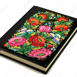 以下が含まれることがあります： 黒い表紙に花柄が描かれたジャーナル。花は赤、ピンク、黄色、青で、対称的なパターンで配置されています。ジャーナルは閉じており、白い表面の上に置かれています。