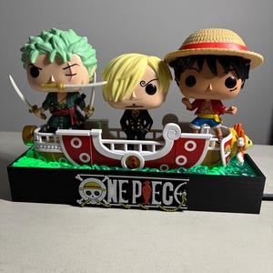 Può includere: Un oggetto da collezione di One Piece con tre personaggi su una base a forma di barca. Le figure includono uno spadaccino dai capelli verdi con spade, un personaggio biondo e un personaggio con un cappello di paglia. La base ha il logo One Piece.