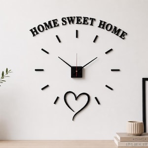 Può includere: Orologio da parete nero con le parole "HOME SWEET HOME" sopra il quadrante. L'orologio ha lancette nere, un centro quadrato nero e indicatori neri. Una forma di cuore nero è sotto l'orologio. Un'immagine incorniciata e un vaso sono anche nell'immagine.