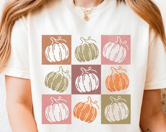 May include: Ivory-colored t-shirt featuring a grid of nine pumpkins in various colors, including orange, green, and pink. The pumpkins are set against a square background. The shirt is a short-sleeved, casual style.