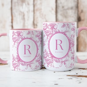 May include: Two white ceramic mugs with pink floral patterns and pink rims. The mugs have a pink circle with the letter "R" and the name "Rachel" in the center.