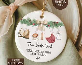 May include: A white ceramic ornament with a gold string for hanging. The ornament features illustrations of books, a coffee cup, and a bow. Text reads "The Book Club" and includes names and the year 2025. The ornament is decorated with green pine needles and red berries.