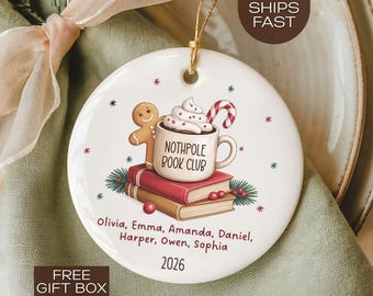 May include: A white ceramic Christmas ornament with a festive design. It features a gingerbread man, a mug with whipped cream, and a candy cane. The text reads "NOTHPOLE BOOK CLUB" and includes names and the year 2026.