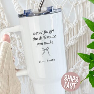 May include: White insulated tumbler with a handle and a black lid. The tumbler has the text "never forget the difference you make" with a bow graphic and "Mrs. Smith" below. A pink sticker with "SHIPS FAST" is visible.
