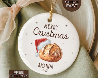 May include: A white ceramic Christmas ornament with the text "MERRY Crustmas" in brown script. A Santa hat and a loaf of bread are illustrated below the text. The ornament also includes the name "AMANDA" and the year "2025".