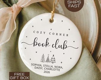 May include: White ceramic ornament with the text "COZY CORNER book club" in black script. The ornament includes the names Sophia, Stella, Nora, Daisy, and Charlotte, with the year 2026. The ornament is decorated with small star designs and two Christmas trees.