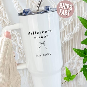 May include: White tumbler with a handle, featuring the text "difference maker" and a bow graphic. Below the bow is the name "Mrs. Smith". The tumbler has a lid and a straw. A pink sticker with "SHIPS FAST" is visible.