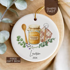 May include: White ceramic ornament with a sourdough starter design. It features a jar labeled "SOURDOUGH STARTER", a wooden spoon, a whisk, and floral accents. The ornament has a gold string for hanging and includes the name "Lauren" and the year "2026".