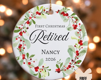 Puede incluir: Un adorno redondo de cerámica blanca con una corona de acuarela de hojas verdes y bayas rojas. El texto dice First Christmas Retired, Nancy y 2026 en letras negras. Un cordón dorado está sujeto en la parte superior para colgarlo.
