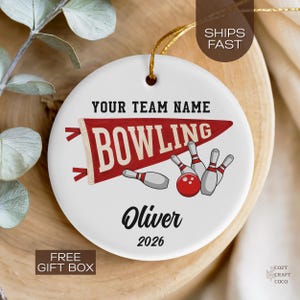May include: A round white ceramic ornament featuring a red pennant flag with the text BOWLING, a red bowling ball, and several white bowling pins. The text YOUR TEAM NAME, the name Oliver, and the year 2026 are printed in black on the ornament.